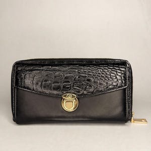 Puede incluir: Una cartera rectangular negra con una sección inferior lisa y una solapa superior con textura de cocodrilo. La cartera se asegura con un cierre giratorio de metal dorado e incluye un tirador de cremallera a juego en el lateral.