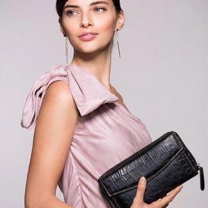 Op de afbeelding: Een lichtroze one-shoulder top met een grote stoffen strik op de schouderband, gecombineerd met een zwarte rechthoekige portemonnee met een krokodillenleer-textuur, ritssluiting en een set goudkleurige metalen armbanden.
