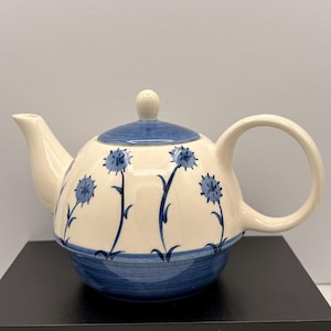 Puede incluir: Una tetera de cerámica de color crema con un patrón floral azul pintado a mano de flores espinosas sobre tallos largos. La tetera incluye una tapa a juego con la parte superior azul, un asa curva, un pico vertedor y una banda azul ancha en la base.
