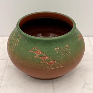 May include: A round ceramic pot with a wide opening, featuring a gradient color scheme that transitions from a deep terracotta brown at the base to a muted forest green at the rim. The exterior surface is decorated with carved geometric symbols.