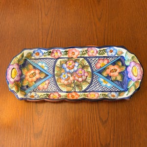 May include: A rectangular ceramic serving tray with scalloped edges, featuring a hand-painted floral pattern in shades of pink, blue, and purple. The design includes a central circular motif surrounded by geometric blue lines and green leaves.