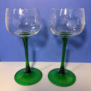 May include: Two clear glass goblets featuring etched floral patterns on the bowls, supported by slender, vibrant green stems and matching circular green bases. The design includes delicate leaf and blossom engravings encircling the upper glass.