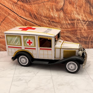 Pode incluir: Um brinquedo de ambulância de metal em estilo vintage com carroceria branca, detalhes dourados, para-lamas pretos e símbolos de cruz vermelha no teto e painéis laterais. O veículo tem janelas com tom amarelo e a palavra AMBULANCE impressa.