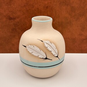 May include: A matte beige ceramic vase with a rounded body and a narrow neck. The vase features two textured white feather designs, a horizontal light blue band encircling the lower section, and a thin black line accenting the blue band.
