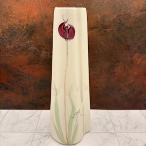May include: A tall, off-white ceramic vase with a tapered, angular silhouette. The front features a hand-painted illustration of a single dark purple flower on a long stem with light green leaves at the base and a signature near the bottom.