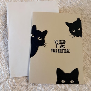 May include: A beige birthday greeting card featuring three black cat silhouettes peeking from the edges. The text in the center reads, "WE HEARD IT WAS YOUR BIRTHDAY." The card is accompanied by a plain white envelope, both displayed on a textured surface.