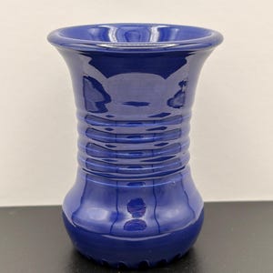 May include: A glossy, deep cobalt blue ceramic vase featuring a wide, flared rim and a cylindrical body with a series of horizontal, raised ridges encircling the neck. The base is rounded and smooth, reflecting light across its polished surface.