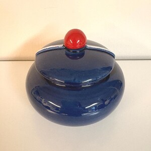 May include: A round, dark blue ceramic jar with a matching lid. The lid features a prominent, glossy red spherical knob at the center and a thin, light blue horizontal band that runs across the top surface, accentuating the circular shape.