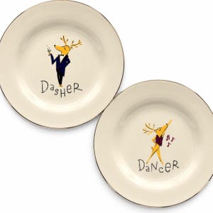 May include: Two cream-colored ceramic plates with metallic rims. One plate features a yellow deer in a dark blue tuxedo holding a cocktail glass with the text "DASHER" below. The other plate shows a yellow deer in a purple vest with musical notes and the text "DANCER" below.