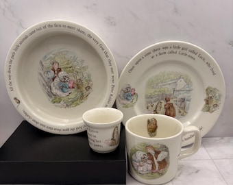 May include: A ceramic dinnerware set featuring two plates, a small cup, and a mug, all decorated with illustrations of characters from Beatrix Potter stories. The cream-colored pieces include printed text from the tales along the rims and sides.