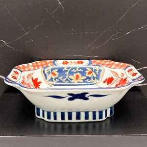 May include: A rectangular ceramic bowl with scalloped edges, featuring a white base decorated with blue and orange floral patterns, a red grid border, and a blue and white striped base. The interior displays blue and orange floral motifs.