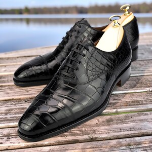 Peut inclure: Une paire de chaussures richelieu noires avec un motif texturé en peau de crocodile brillant. Les chaussures ont des lacets noirs, un bout arrondi et sont équipées d'embauchoirs en bois clair avec des boutons en métal doré au talon.