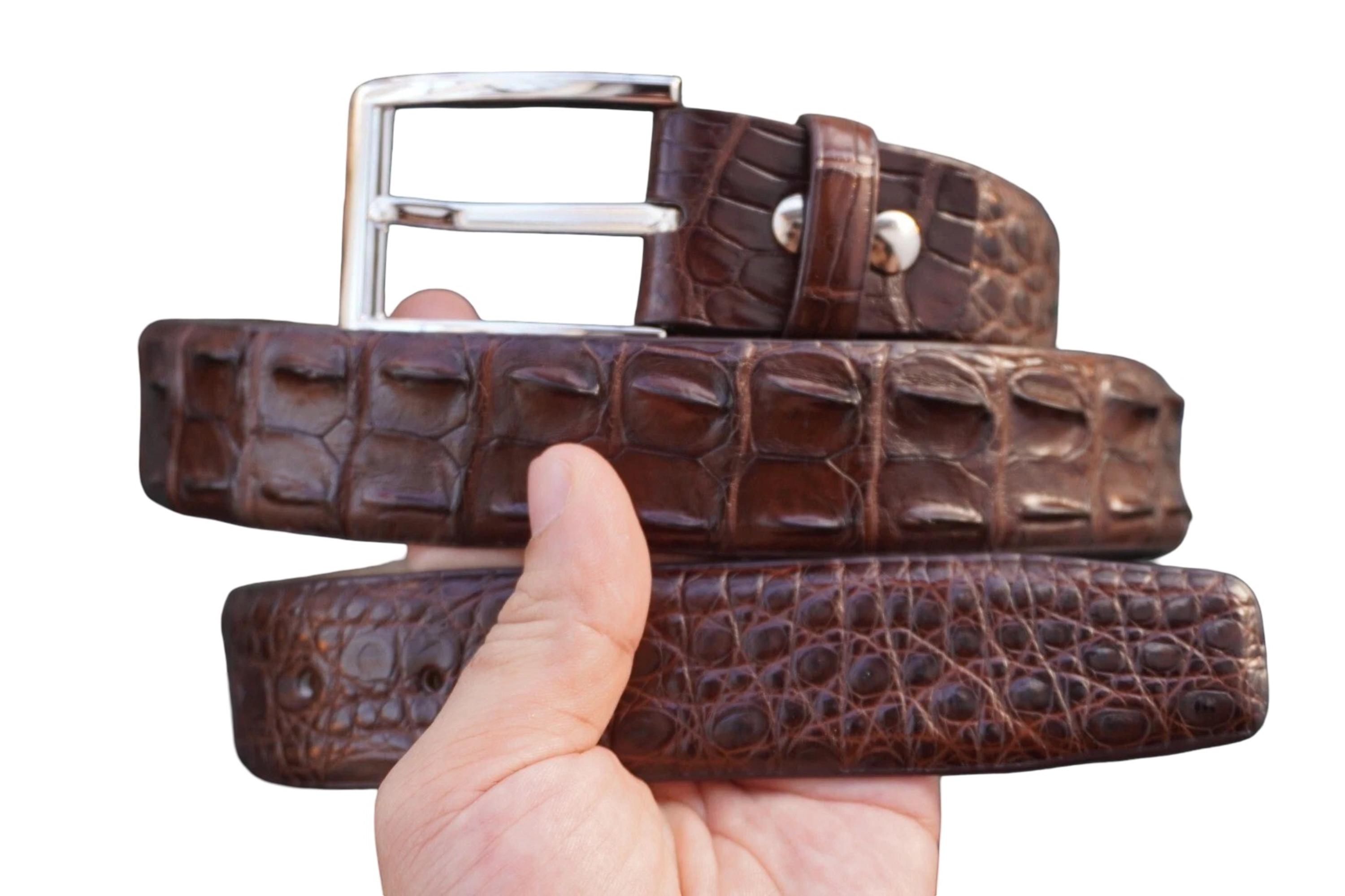 Pode incluir: Um cinto castanho escuro com um padrão texturizado e em relevo que lembra pele de crocodilo. O cinto inclui uma fivela de metal prateado polido com uma estrutura retangular e um pino, fixada por dois rebites prateados perto da fivela.