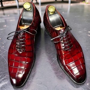 Peut inclure: Une paire de chaussures oxford rouge foncé et brillantes avec un motif texturé en peau de crocodile. Chaque chaussure possède des lacets noirs, un bout pointu et un embauchoir en métal doré inséré pour maintenir la forme de la chaussure.