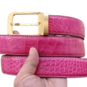 Pode incluir: Um cinto rosa brilhante com um padrão de textura de pele de crocodilo e uma fivela de metal dourado polido. O cinto está enrolado em um laço em camadas, exibindo a cor fúcsia vibrante e a superfície detalhada em relevo em toda a tira.