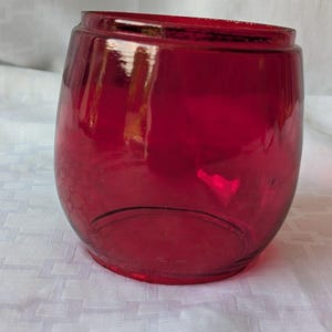 Könnte beinhalten: Ein tiefroter Votivkerzenhalter aus rubinrotem Glas mit einer abgerundeten, fassartigen Form. Das Glas ist durchscheinend und glatt, mit einer breiten kreisförmigen Öffnung an der Oberseite und einer etwas schmaleren Basis, was eine einfache, geschwungene Silhouette ergibt.