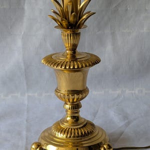 Puede incluir: Una lámpara de mesa de latón pulido con un cuerpo central en forma de urna con detalles estriados, un remate decorativo en forma de piña en la parte superior y una base circular sostenida por tres pequeños pies curvos. Se ve un casquillo.