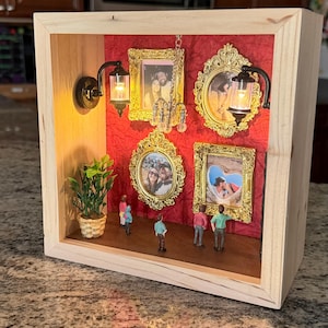 May include: A wooden shadow box display featuring a red textured background with four ornate gold frames containing photographs. The scene includes two miniature wall-mounted lamps, a small hanging chandelier, a potted plant, and five small figurines.
