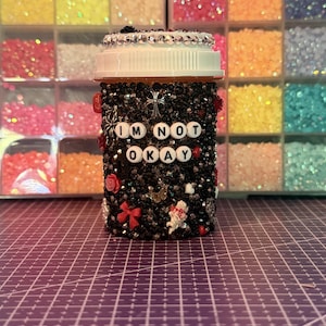 May include: A cylindrical container covered in black rhinestones, featuring white letter beads spelling "I'M NOT OKAY" on the front. The lid is rimmed with silver rhinestones, and the body is decorated with small charms including a cross, red bows, and roses.