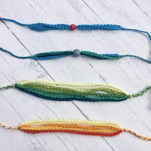 May include: Four handmade bracelets made of braided thread. The top two are blue with single beads in pink and gray. The third is a multi-strand bracelet in shades of green and yellow. The bottom bracelet features orange and yellow threads.