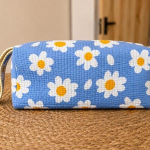 Könnte beinhalten: Eine rechteckige Reißverschlusstasche aus blauem Steppstoff mit einem sich wiederholenden Muster aus weißen und gelben Gänseblümchen. Die Tasche verfügt über einen seitlichen Tragegriff aus gelb-weiß kariertem Stoff und eine texturierte Gitteroberfläche.