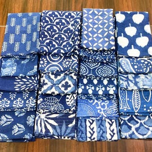 May include: A collection of sixteen folded square fabric napkins arranged in a four-by-four grid. Each napkin features a distinct blue and white block-printed pattern, including botanical motifs, geometric shapes, and abstract floral designs.