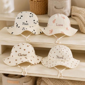Op de afbeelding: Vier crèmekleurige bucket hats met kinbandjes. De hoed linksboven heeft zwarte pandagezichten en de naam Finn. De hoed rechtsboven heeft roze konijnenmotieven en de naam Luna. De hoed linksonder heeft bruine hartjes en de naam Carter. De hoed rechtsonder heeft bruine stippen en de naam Nora.