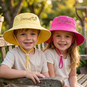 May include: A yellow bucket hat with the name Samuel and a raccoon icon embroidered on the front, and a pink bucket hat with the name Sophia and a rabbit icon embroidered on the front. Both hats feature adjustable chin straps with toggles.