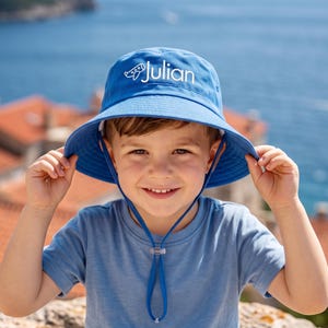 May include: A blue bucket hat featuring a white embroidered graphic of a small airplane and the name Julian in white text. The hat includes a matching blue chin strap with an adjustable plastic toggle for a secure fit around the head.