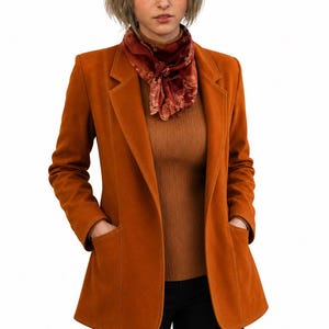 Op de afbeelding: Een roestoranje blazer met een ingekeepte revers en twee opgestikte voorzakken, gedragen over een bruine geribbelde coltrui en een donkerrode sjaal met bloemenpatroon, gecombineerd met een zwarte broek, met een zachte suède-achtige textuur.