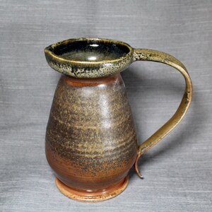 May include: A brown ceramic pitcher with a speckled glaze. The pitcher has a wide, rounded base and a narrow neck. The handle is attached to the side of the pitcher and curves up and over the top.