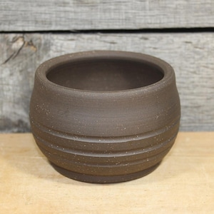 May include: A small, round, brown ceramic pot with a textured surface and horizontal grooves around the base. The pot has a wide opening and sits on a flat, circular base. The background is a wooden surface.