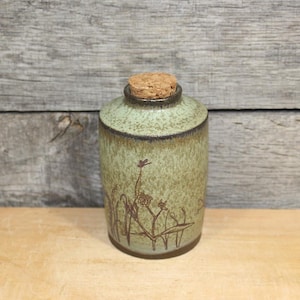 May include: A small, cylindrical ceramic jar with a cork stopper. The jar has a light green glaze with brown floral designs. The rim and base are a darker brown. The jar is approximately 4 inches tall.