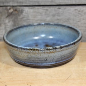May include: A round ceramic bowl featuring a mottled blue and grey glaze with a dark, speckled rim. The interior of the bowl displays a smooth, concave surface with subtle variations in color, while the exterior has a textured, earthy finish.