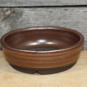 May include: A round, shallow ceramic bowl with a dark brown, metallic-toned glaze. The exterior features a subtle horizontal ridge detail near the rim, while the base is a contrasting matte dark brown color with a small notch cut into the bottom edge.