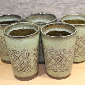 May include: Five ceramic tumblers with a light green glaze and a brown Celtic knot design. The tumblers are filled with a dark green substance. The rims have a darker brown color. The tumblers are on a wooden surface.