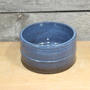 May include: A handmade, round ceramic bowl with a speckled blue glaze. The bowl has a dark brown base and two horizontal grooves around the middle. The bowl is sitting on a light-colored wooden surface.
