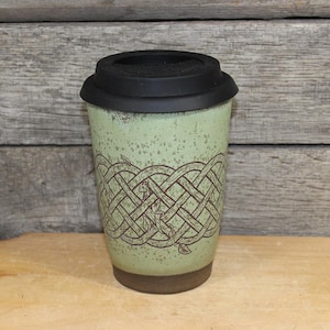 May include: A ceramic travel mug with a black lid and a light green body. The body features a brown Celtic knot design. The mug has a brown base and is designed to hold hot or cold beverages.