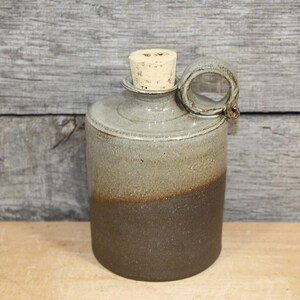 May include: A cylindrical ceramic bottle featuring a two-tone glaze with a light gray upper section and a dark brown lower section. The bottle has a narrow neck, a single circular side handle, and is sealed with a light-colored cylindrical cork stopper.