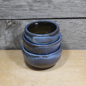 May include: A stack of three handmade ceramic bowls. The bowls are glazed in shades of blue and brown, with a textured surface. The bowls are stacked on top of each other, with the largest bowl at the bottom and the smallest at the top. The bowls are on a wooden surface.