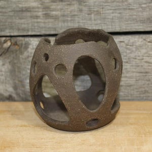 May include: A brown, spherical ceramic candle holder with multiple oval and circular cutouts. The decorative piece is made of textured, speckled clay and sits on a wooden surface. The background features a weathered wooden wall.