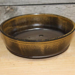 May include: A round, shallow ceramic bonsai pot with a dark brown and golden-yellow mottled glaze. The exterior features a smooth, slightly tapered wall with a defined rim, while the interior base contains visible drainage holes for plant care.