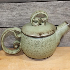 May include: A handcrafted, speckled green ceramic teapot with a matching lid. The teapot features a curved handle and spout, and a decorative swirl on the lid. The teapot is a unique piece of pottery.