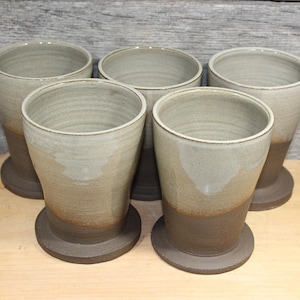 May include: Five handmade ceramic tumblers. Each tumbler has a light beige upper half and a dark brown lower half, with a matching dark brown base. The tumblers are cylindrical with a slightly flared top.