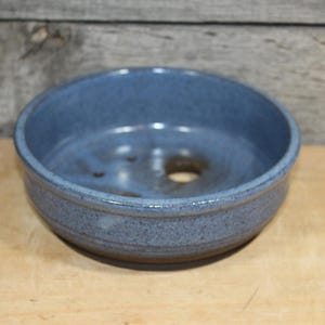 May include: A round, shallow ceramic bowl with a speckled blue glaze. The interior features a smooth, glossy finish with a visible drainage hole at the bottom, while the exterior displays a slightly darker, textured blue tone with a rounded rim.