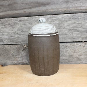 May include: A handmade ceramic jar with a lid. The jar is a dark brown color with vertical textured lines. The lid is white with horizontal lines and a small knob on top. The jar is sitting on a wooden surface.