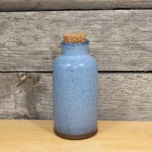 May include: A small, speckled blue ceramic bottle with a cork stopper. The bottle has a rounded body and a narrow neck. The base of the bottle is a darker brown color. The background is a wooden surface.