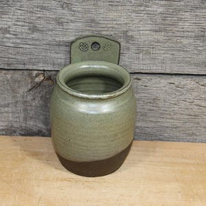 May include: A wall-mounted ceramic vessel with a rounded body, featuring a sage green glazed upper section and a dark brown unglazed base. The back plate includes a circular hole for hanging and two small stamped leaf motifs on either side.