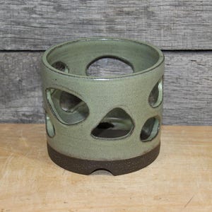May include: A cylindrical ceramic candle holder in a muted green hue, featuring irregular cut-out shapes. The base is a darker brown, and the piece sits on a wooden surface. This decorative item is suitable for home decor.