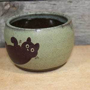 May include: A round ceramic bowl with a speckled light green exterior and a dark brown interior. The outer surface features a dark brown graphic illustration of a cat lying on its back with its paws raised and wide eyes looking forward.